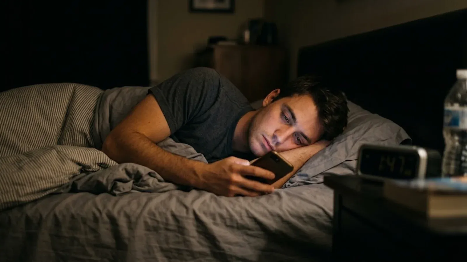 A young man lying in bed late at night scrolling on his phone in a dark bedroom