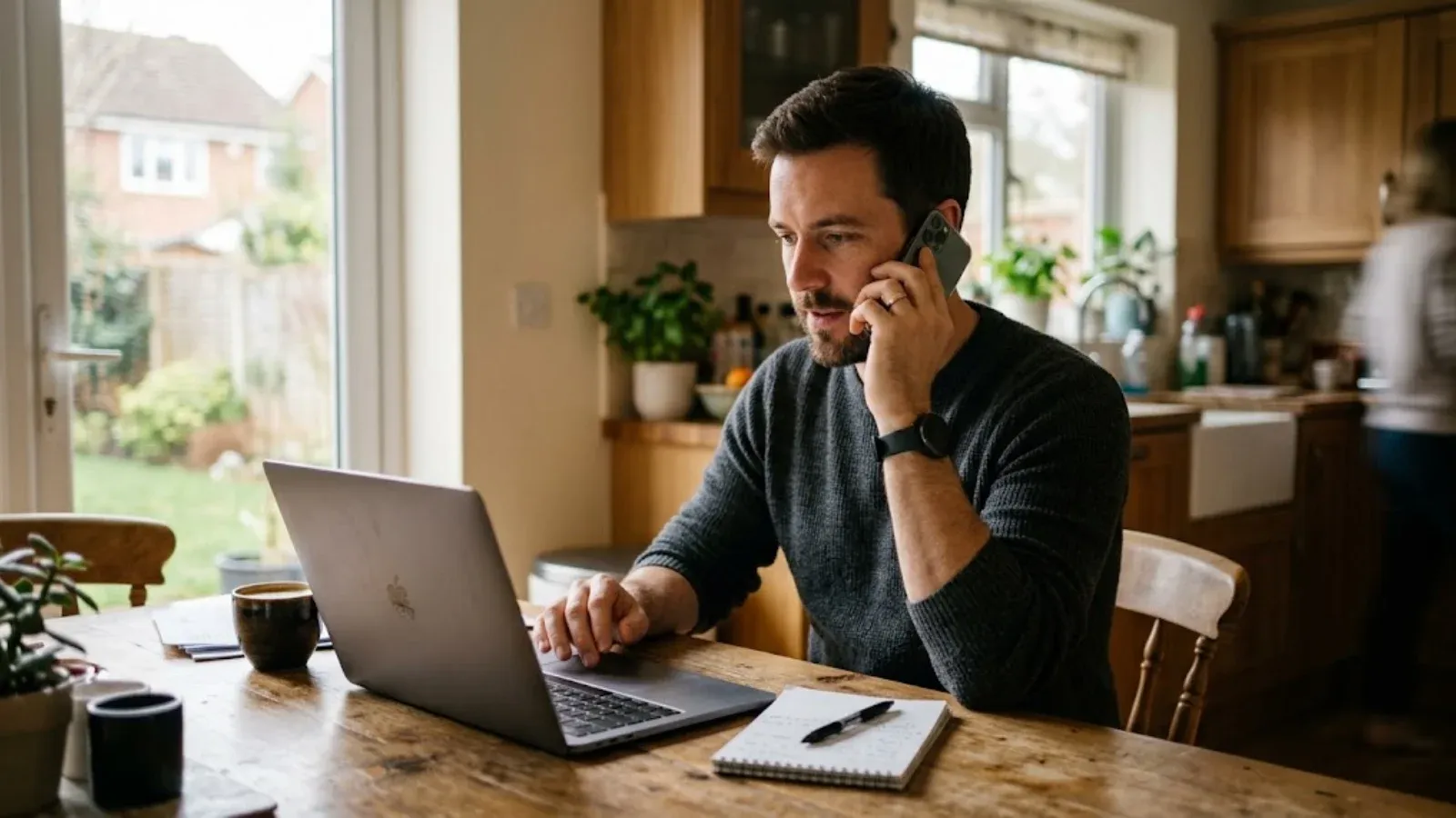 A man on a phone call at his kitchen table checking details on a laptop